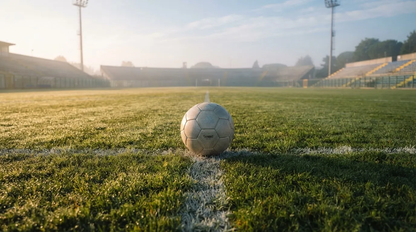 Ein Fußball liegt auf dem Mittelkreis eines leeren Rasenplatzes am frühen Morgen, im Hintergrund eine ferne Tribüne im Dunst.