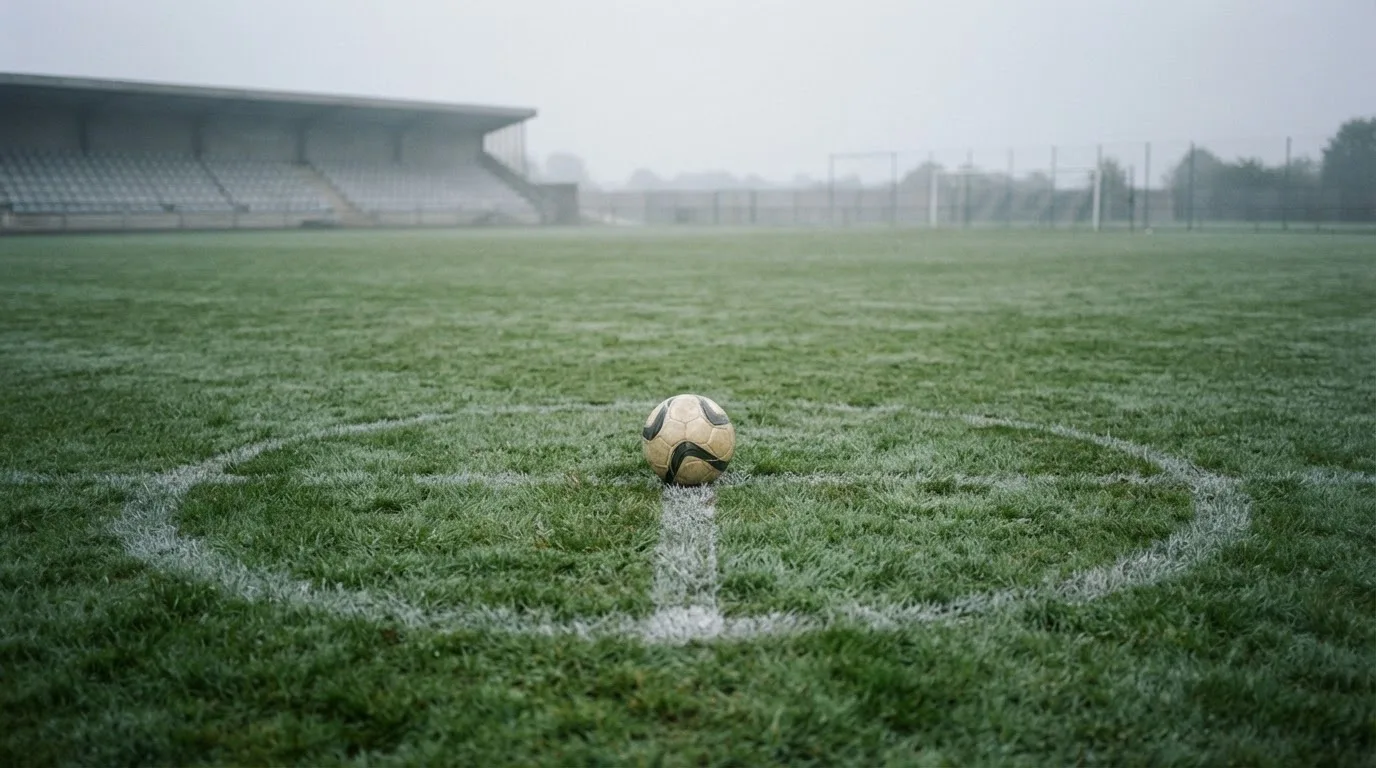 Ein Fußball liegt auf dem Mittelkreis eines leeren Rasenplatzes am frühen Morgen, im Hintergrund eine ferne Tribüne im Dunst.