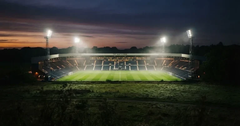 Ein Stadion unter Flutlicht aus der Ferne am Abend, hell erleuchtet, Nebel liegt über dem Rasen.