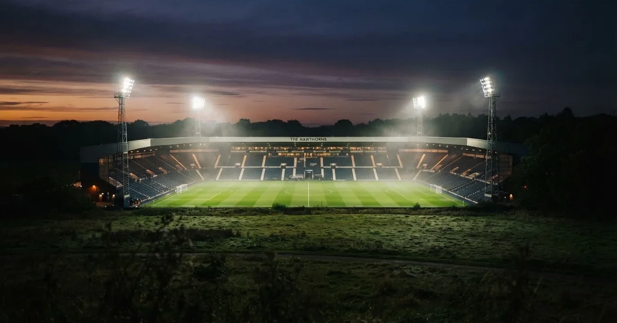 Ein Stadion unter Flutlicht aus der Ferne am Abend, hell erleuchtet, Nebel liegt über dem Rasen.