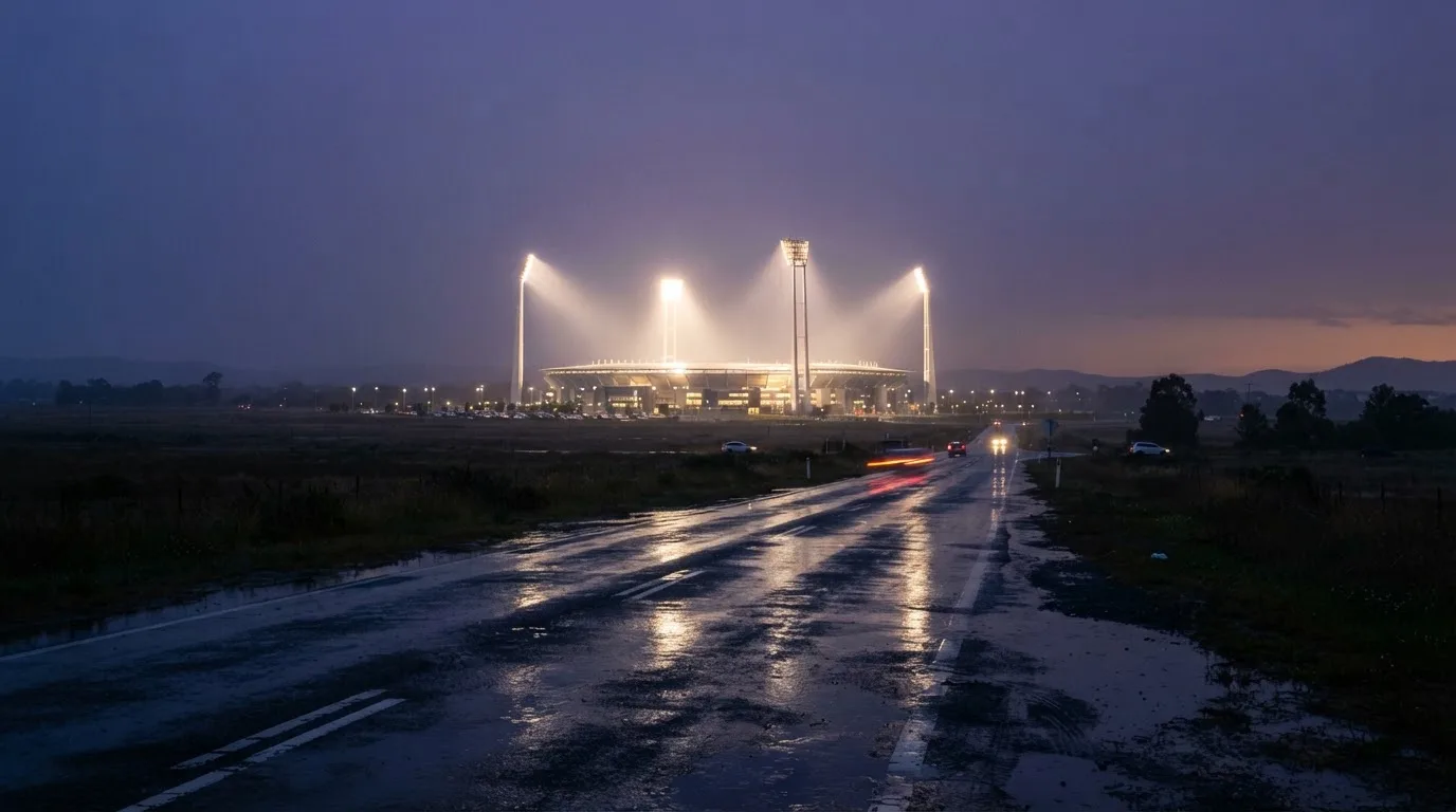 Ein Stadion unter Flutlicht aus großer Entfernung gesehen, davor eine nasse Straße in der Abenddämmerung.