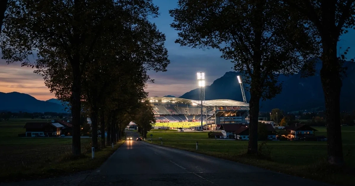 Ein Stadion unter Flutlicht aus großer Entfernung gesehen, davor eine dunkle Landstraße und einzelne Bäume in der Abenddämmerung.