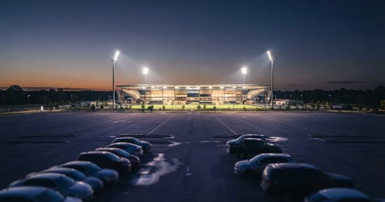Ein Fußballstadion unter Flutlicht aus großer Entfernung gesehen, die Lichtkegel stehen im dunklen Abendhimmel über leeren Parkplätzen.