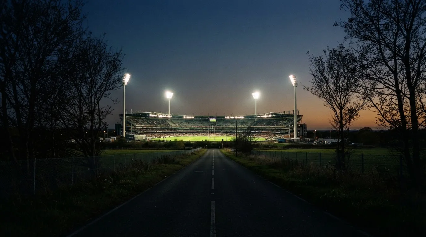 Ein großes Stadion unter Flutlicht aus weiter Entfernung gesehen, davor eine dunkle Landstraße und ein paar Bäume.