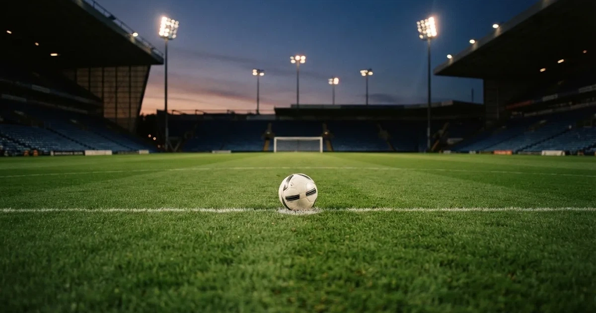 Ein Fußball liegt auf dem Anstoßpunkt eines Rasenplatzes in der Dämmerung, in der Ferne leuchten Flutlichter, die Ränge sind leer.